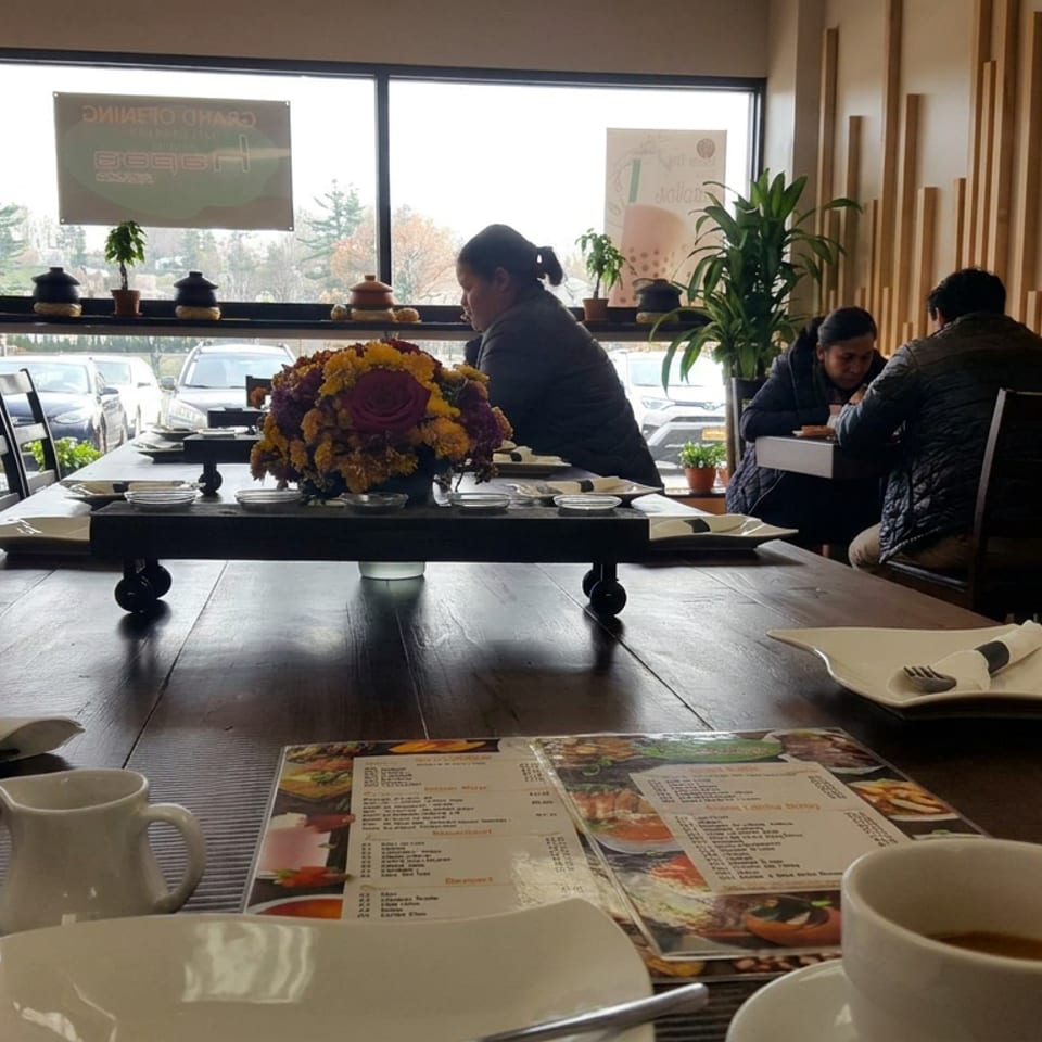 Warmly lit restaurant dining room with set tables, floral centerpiece, menus, plants, and diners seated near large windows.