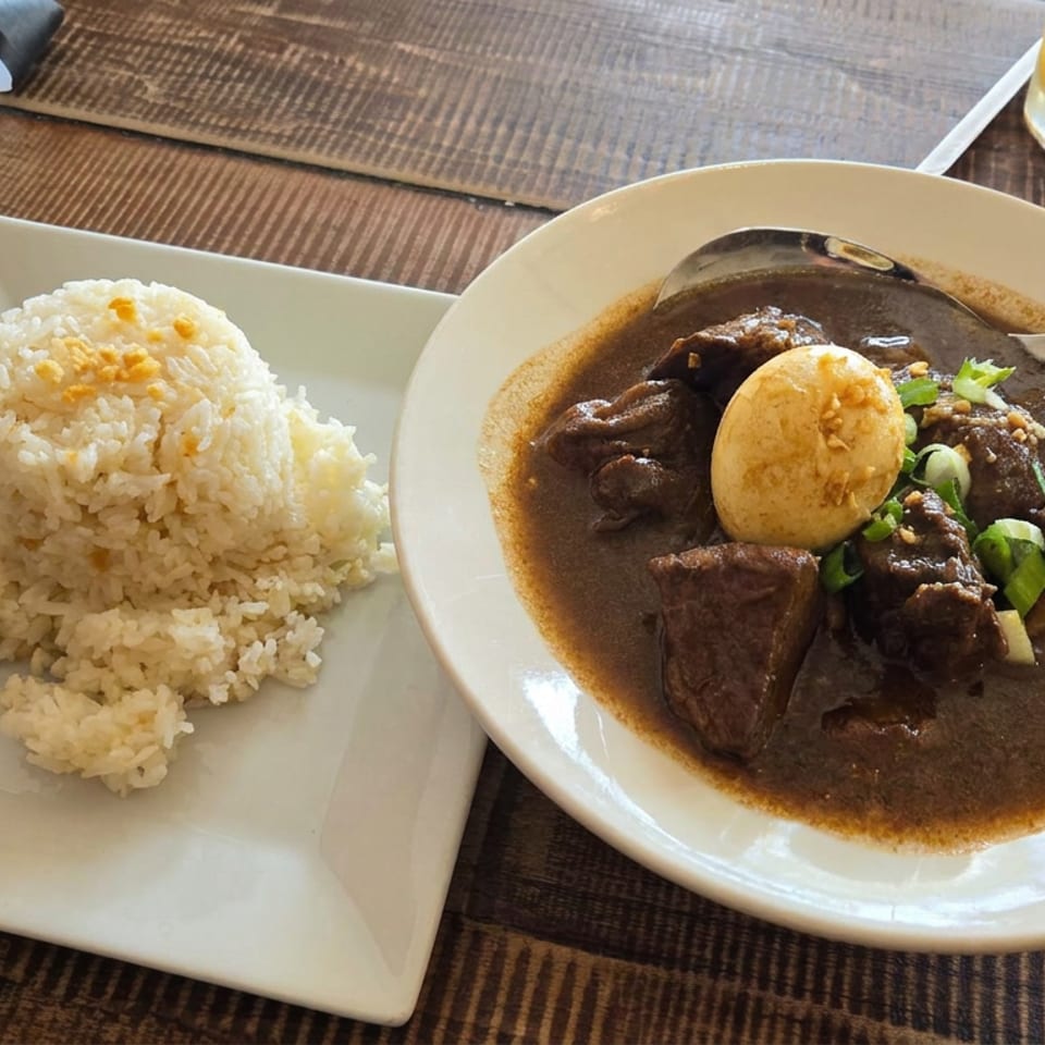 Plate of beef stew with boiled egg and green onions beside a mound of white rice on a wooden restaurant table.