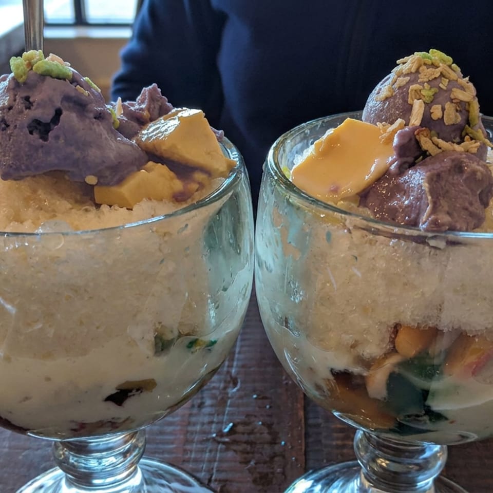 Two glass dessert bowls filled with shaved ice, ice cream, fruit, sweet toppings, and crushed nuts on a restaurant table.