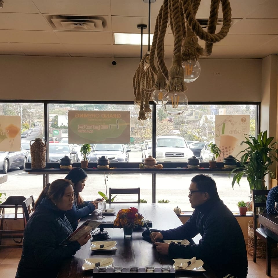 Bright restaurant interior with diners at wooden tables, rope pendant lights, potted plants, and large windows overlooking parked cars.